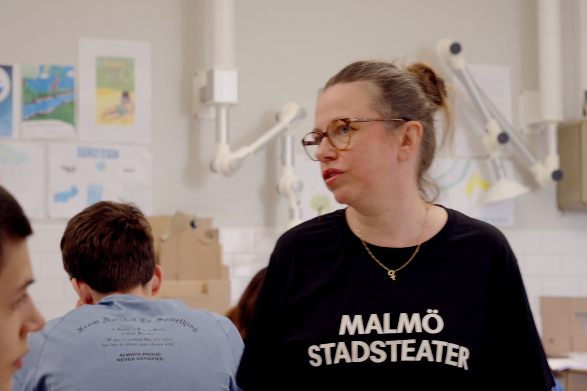 Hedvig i Malmö Stadsteater tshirt, runda glasögon, uppsatt hår i knut och guldhalsband med kvinnosymbol pratar med unga som skymtar i bild.