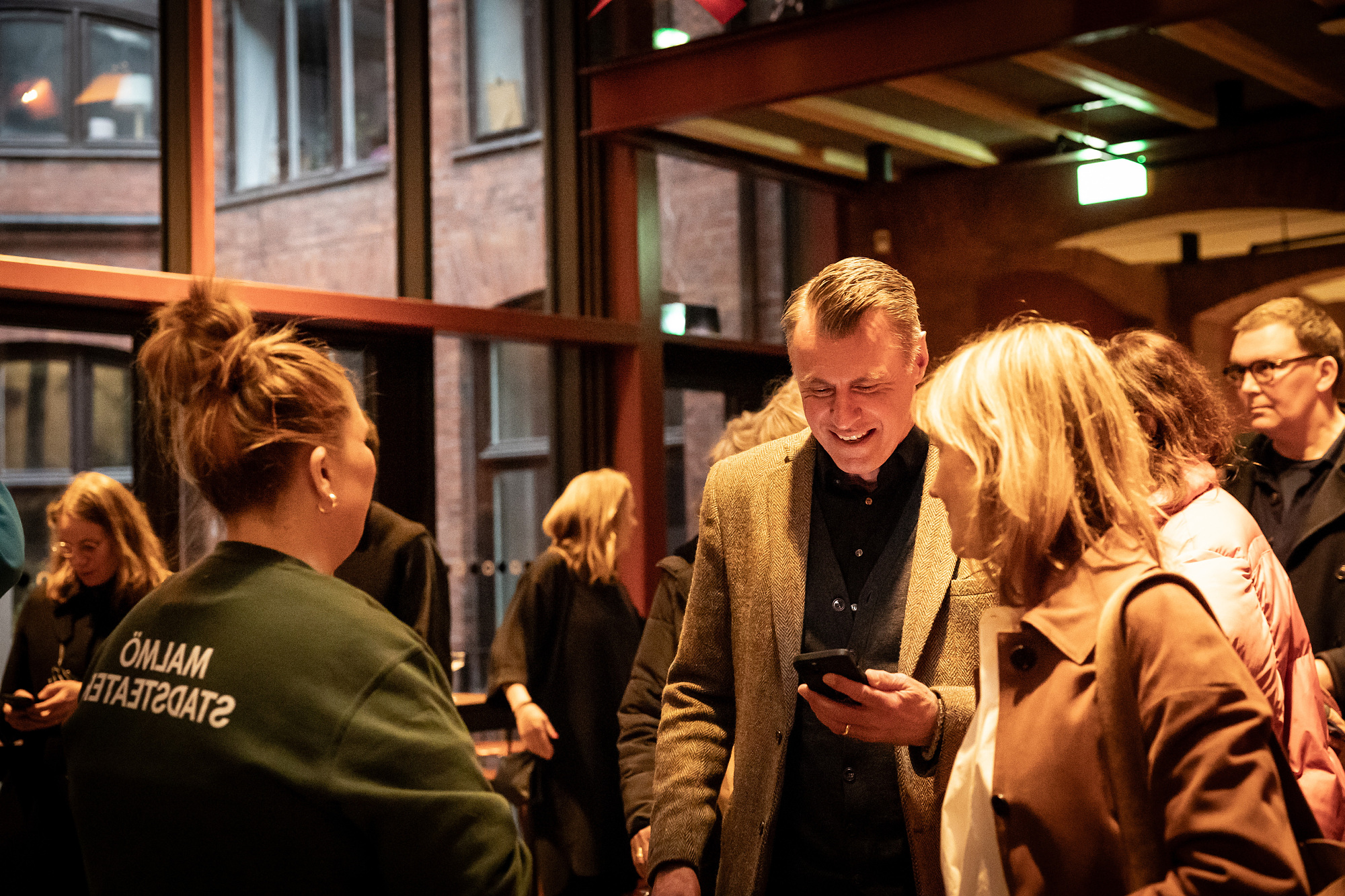 En publikvärd i grön Malmö Stadsteater-jacka hjälper gäster under premiärminglet, medan en man i kavaj ler och tittar på sin mobil.