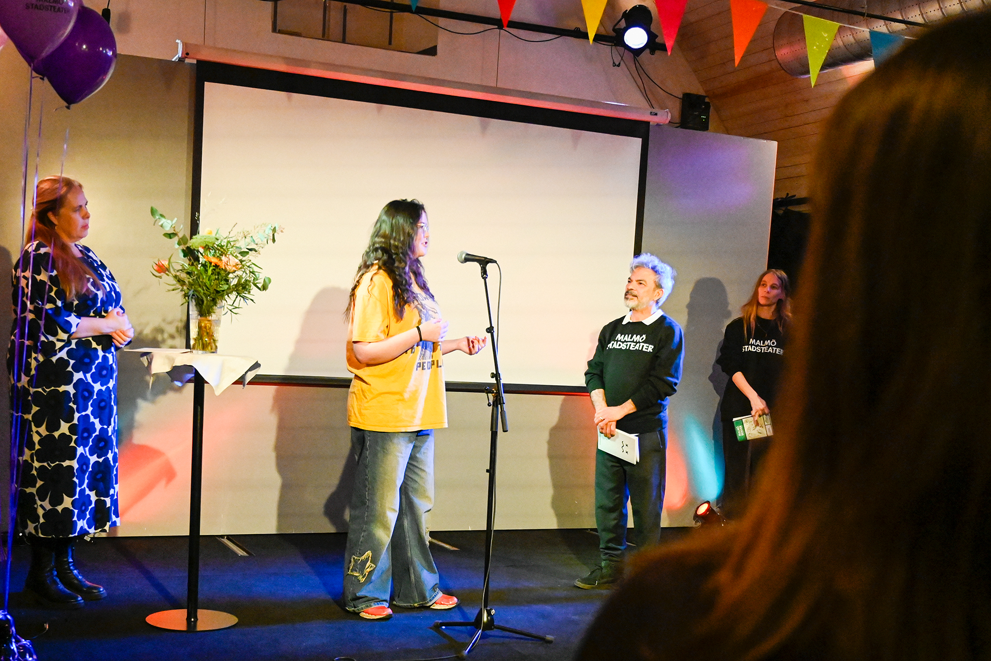 En ung kvinna i gul t-shirt pratar i en mikrofon på en scen medan en man i svart tröja med texten "Malmö Stadsteater" lyssnar. Färgade vimplar och ballonger dekorerar lokalen.