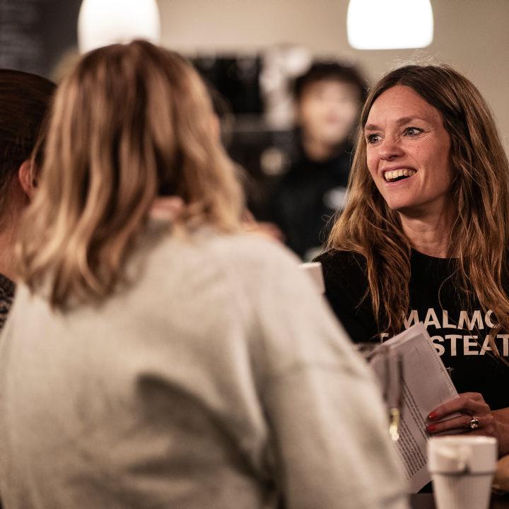 En person med Malmö Stadsteatertröja syns prata med två personer som har ryggen mot kameran.