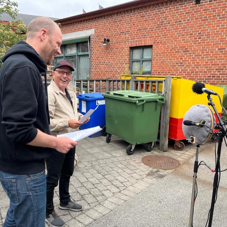 Två personer står på en bakgård bredvid en soptunna och framför en mikrofon.