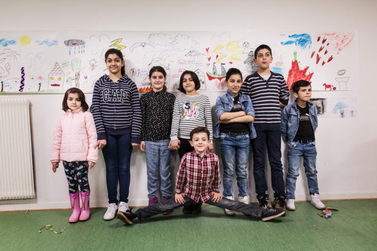 Åtta barn står framför en lång rad teckningar uppsatta på en vägg. De ser stolta och glada ut.