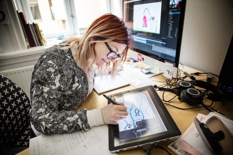 Kvinna sitter vid skrivbord och tecknar på en Ipad bredvid en datorskärm.