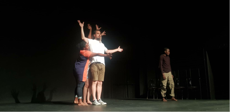 Bild med fyra personer som står på en scen och spelar teater. En kvinna kramar en man som står och öppnar armar. 
