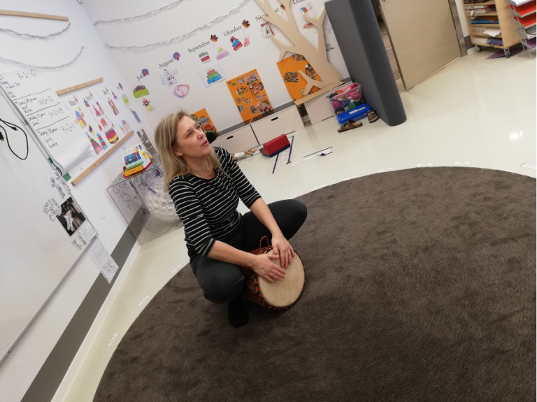 en kvinna sitter på en matta i ett klassrum och håller en trumma med händerna. 