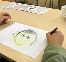 Ett barn sitter vid ett bord och ritar en teckning av ett ansikte på ett vitt papper med färgpennor. Ansiktet är färglagt med gula och svarta toner och har stora blå ögon. På bordet syns en korg med pennor och papper med alfabetet och pedagogiska bilder. Barnets armar är klädda i en grön tröja och i handen håller barnet en mörk penna.